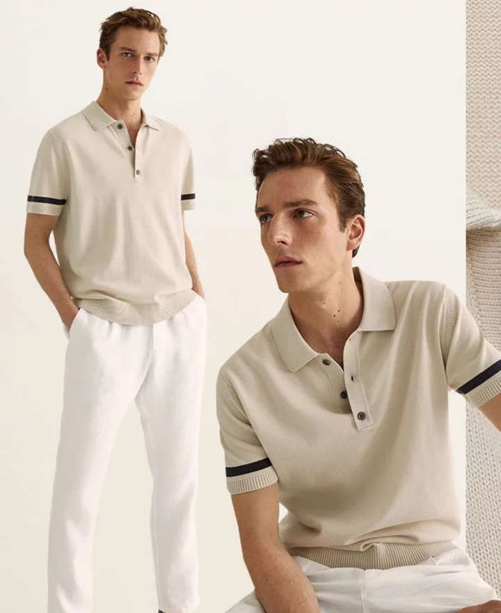 Two male models wearing a light beige short-sleeve knit polo shirt with black stripe accents on the sleeves, paired with white trousers, set against a clean off-white background.